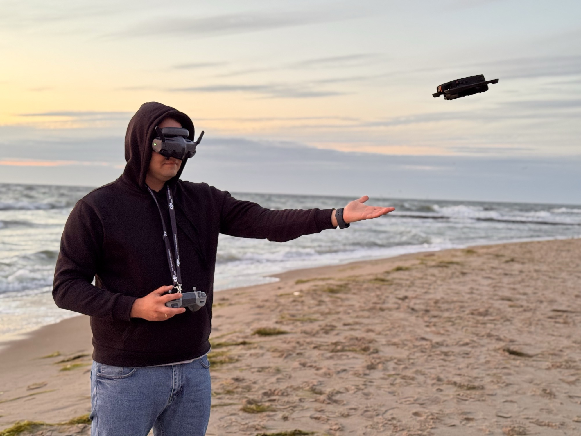 Paweł Worwa w goglach FPV, dron ląduje mu na dłoni na plaży o zachodzie słońca